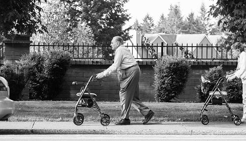 Photo of an old man and woman, each pushing a wheeled walker, strolling down a sidewalk.