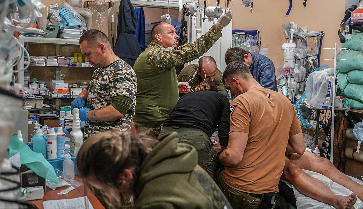 A soldier is treated in a small room crowded with medics and supplies.