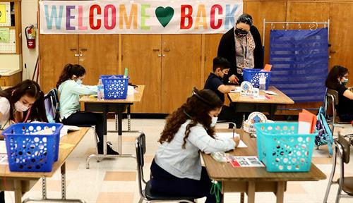 Elementary school students wear face masks while they sit at their own desks in a classroom in Los Angeles in April 2021. Their masked teacher walks amongst them, with a Welcome Back sign on the wall behind.