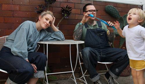 Photograph of a woman and a man sitting at a table in a courtyard while a young child jumps in the air. The man is holding a bubble blower. The woman is leaning on the table, staring ahead.