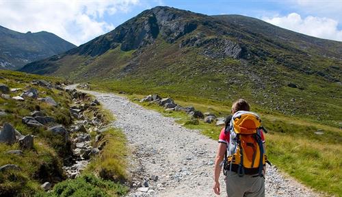 Photograph of a person wearing gray pants and tee shirt heading up a rocky trail. The person is wearing a backpack. There are bleak hills ahead, with low vegetation.