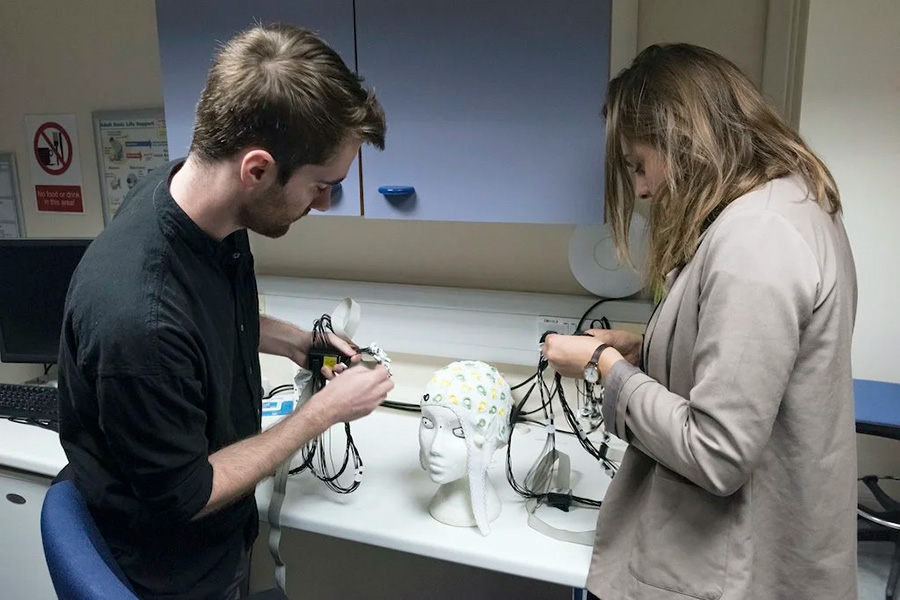 Photograph of two individuals working with electronic equipment on a lab bench. They are leaning over a model of a human head upon which a cap with electrodes is placed.
