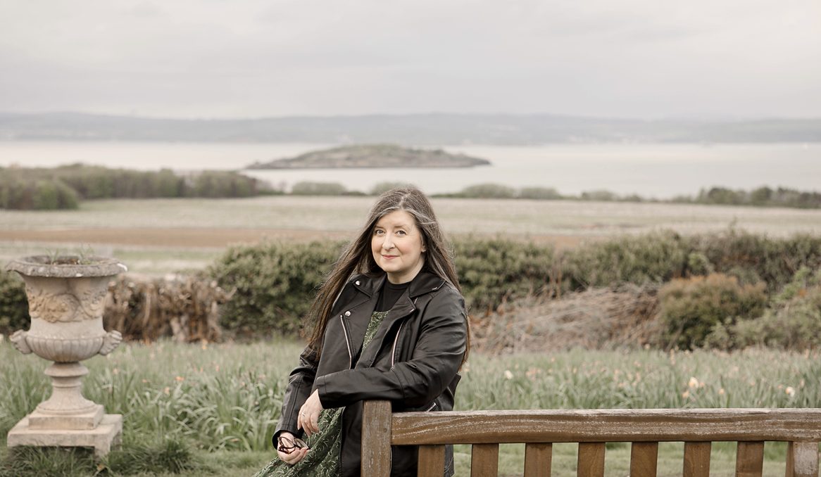 Photograph of a woman with long hair standing in the countryside with a large body of water behind her.