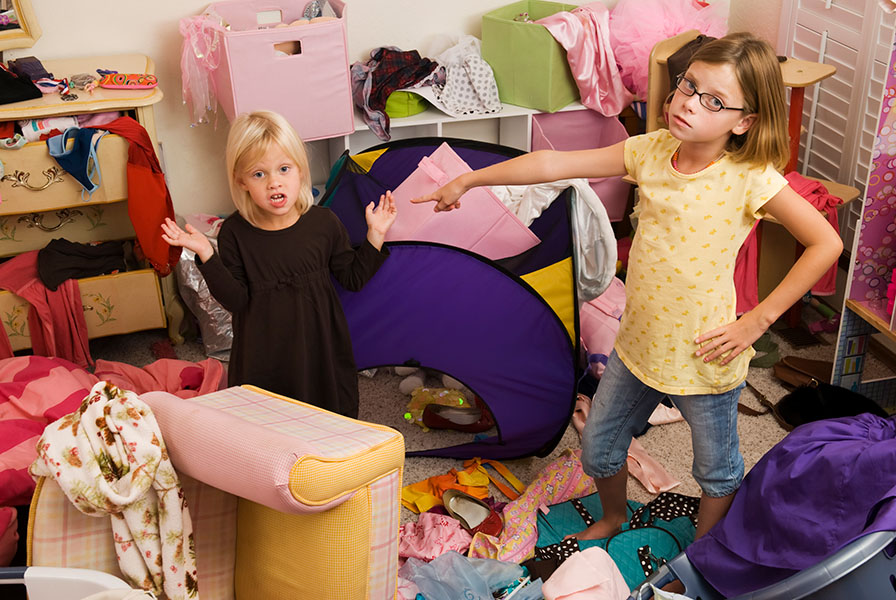 Two young girls in a bedroom, one pointing at the other in an accusing gesture.