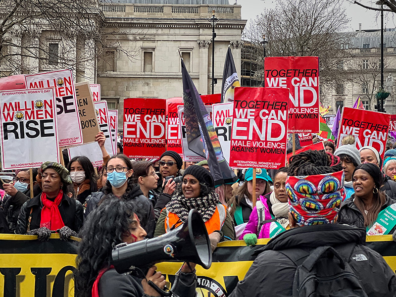 A crowd of women march in London carrying placards that read “Million Women Rise” and “We can end male violence against women.” A woman carries a bullhorn in the foreground.