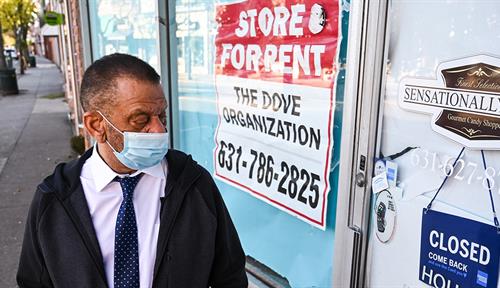 Photograph of a man wearing a surgical mask standing on a sidewalk — he is staring at the door of a store called “Sensationally Sweet.” The windows and glass of the door are covered on the inside with paper. There is a large “Store for rent” sign in the window.