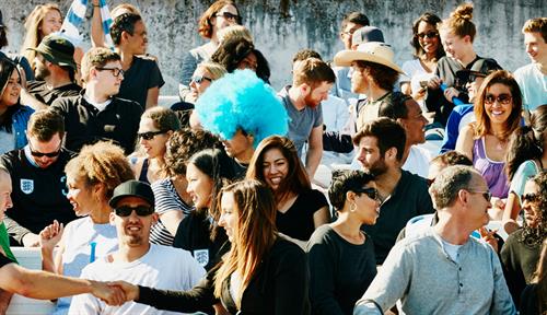 Photograph of several people sitting in bleachers in a stadium taking part in many kinds of social interactions, such as shaking hands, kissing and talking.