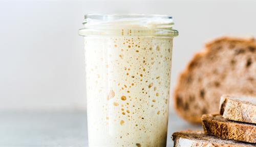 A foaming jar of sourdough starter stands next to a sliced loaf of bread.