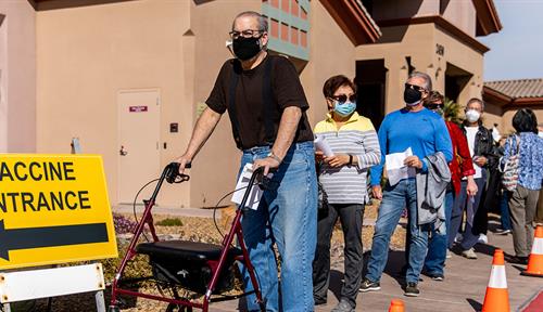 Photograph of a line of people standing on a sidewalk wearing masks. A large yellow sign with an arrow reads “Vaccine entrance.” Orange cones mark one side of the sidewalk, and there is a building along the other side.